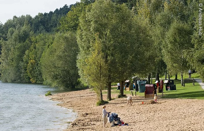 Eine Familie erkundet den großzügigen  Strand des Igelsbachsees, der den Kindern jede Menge Platz zum Spielen bietet. Die Strandkörbe und Bäume bieten Schutz vor der Sonne.