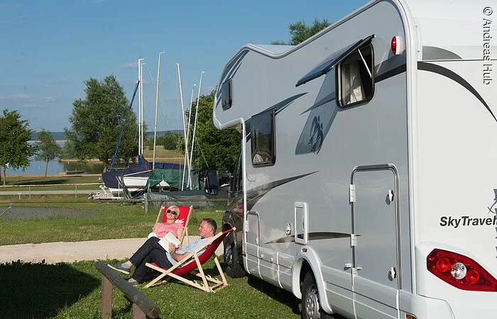 Eine Frau und ein Mann sitzen in zwei Liegestützen link neben einem Wohnmobil. Im Hintergund lagern Boote auf Anhängern. Dahinter beginnt der See.