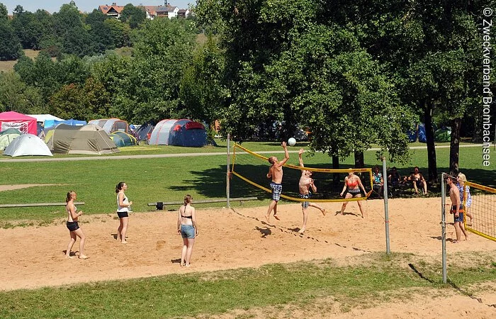 Junge Leute bespielen eines der Beachvolleyball-Felder auf der Badehalbinsel Absberg. Im Hintergund sind grüne Bäume und ein Zeltplatz mit bunten Zelten zu erkennen.