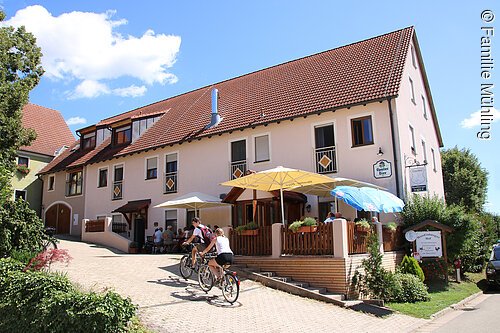 Gastwirtschaft "Noudlsberger Hof" Seitansicht der Gastwirtschaft Noudlsberger Hof. Ein rosa farbenes Haus mit einer Terasse mit Sitzgelegenheiten und Sonnenschirmen direkt davor. Zwei Radler radeln die Einfahrt des Hofes hoch.