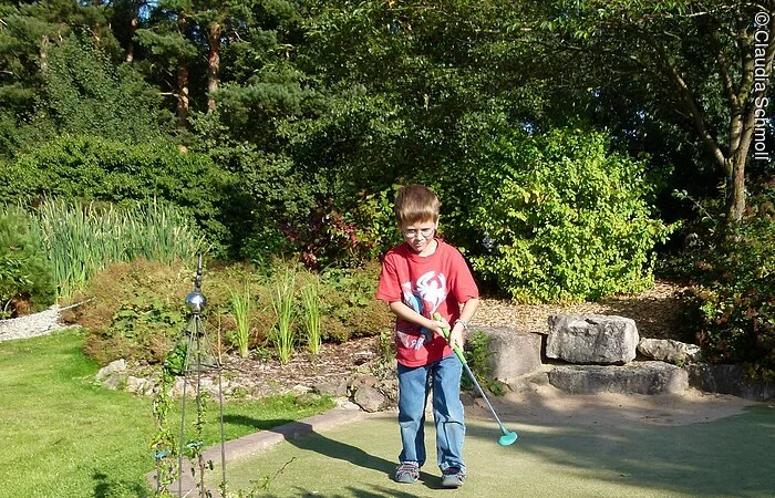 Ein Junge spielt erfreut Minigolf in Pfofeld auf der Minigolfanlage "Amigo" bei strahlendem Sonnenschein und umgeben von grünen Strüchern und Bäumen.