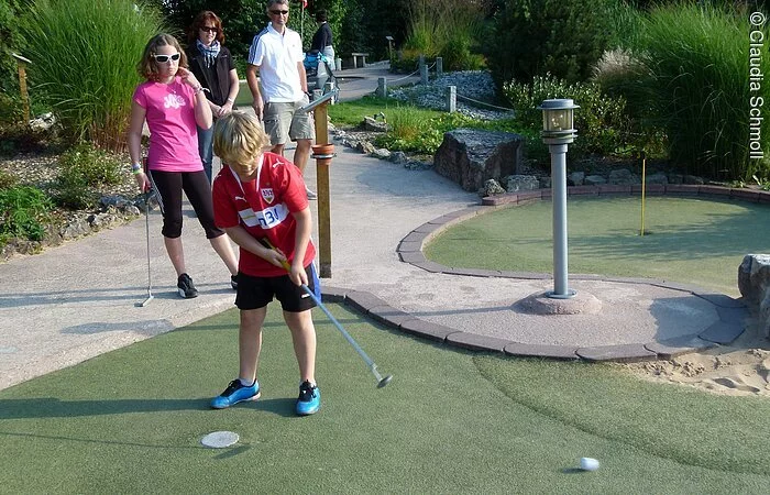 Eine Familie macht einen kleinen Ausflug zum Minigolfplatz "Amigo" in Pfofeld und spielt um die Wette. Der Sohn ist gerade an der Reihe und steht auf dem grünen Platz umgeben von grünen Sträuchern und Bäumen.