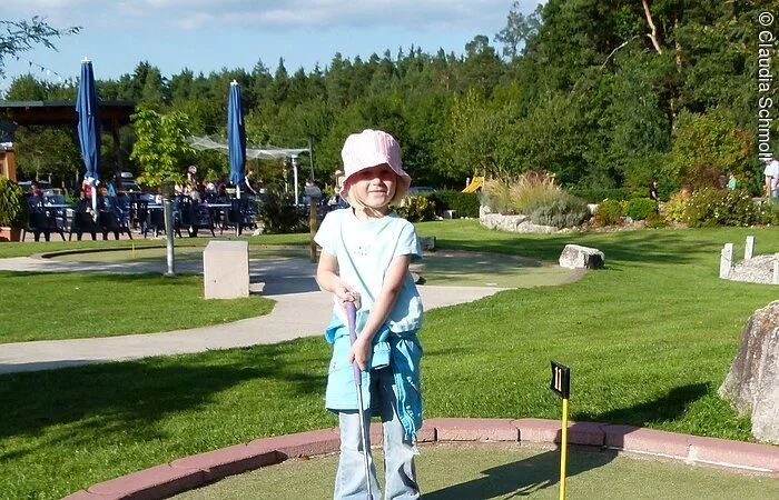 Auf dem Minigolfplatz "Amigo" in Pfofeld spielt ein kleines Mädchen Minigolf und versucht gerade einen Ball einzulochen. Hinter ihr sitzen Gäste im Biergarten und genießen die Sonne.