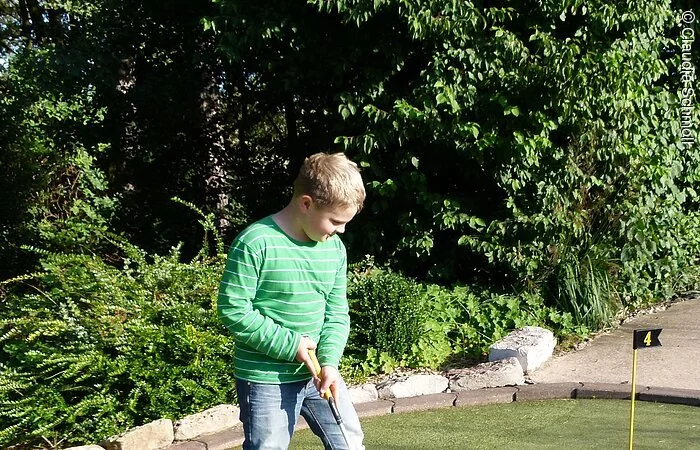 Auf dem Minigolfplatz "Amigo" in Pfofeld spielt ein Junge begeistert Minigolf. Er steht auf einem grünen PLatz und hinter ihm grüne Bäume und Sträucher.