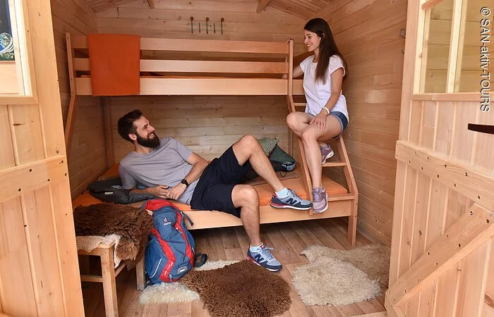 Ein Pärchen sitzt lächelnd auf einem Stockbett in einem kleinen holzverkleideten Schlafraum.