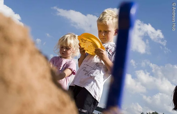 Blick auf zwei Kinder die im Sand spielen. Dahinter blauer Himmel. Im Vordergrund ist verschwommen eine Schaufel und ein Sandhaufen zu erkennen.