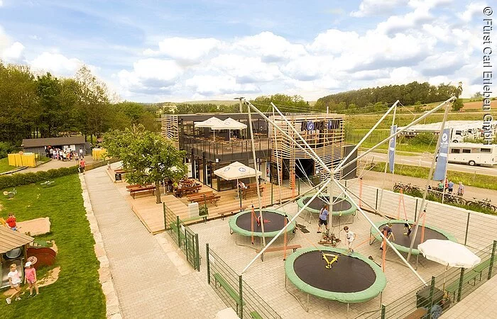 Ein großes Holzhaus mit kleinem Biergarten. Davor vier große Trampoline. Das Grundstück ist umgeben von grüner Wiese.