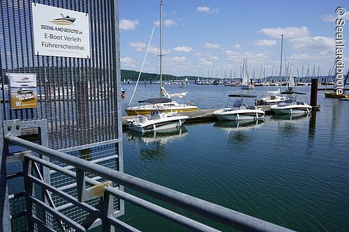 Blick auf den Bootsverleih direkt am Ramsberger Yachthafen. Links im Bild ein Gitter auf welchem ein Schild befestigt ist: Seenlandboote E-Boot Verleih Führerschein frei