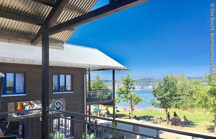 Blick von einem Balkon auf ein zweistöckiges Holzhaus mit Terasse. Dahinter eine Liegewiese mit See.