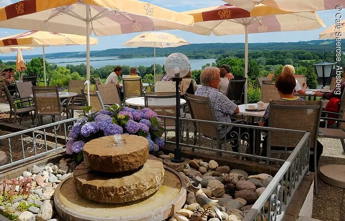 Im Vordergrund ein kleiner Springbrunnen aus Stein. Dahinter sitzen Gäste des Café Seerose auf der Terasse mit Blick auf den See. Auf der Terasse stehen viele Tische mit Stühlen sowie orangene Sonnenschirme.