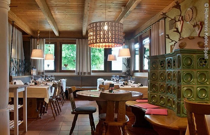 Gastraum mit Stühlen und Tischen aus Holz. Im rechten Bildrand ein Kachelofen mit grünen Kacheln und einer Holzbank an welcher ein runder Tisch steht. Die Decke ist aus Holz, der Boden gefließt.