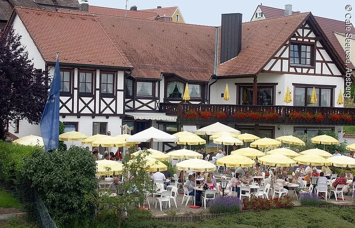 Außenansicht des Cafes "Seerose" mit gemütlicher Terasse in Absberg am Kleinen Brombachsee. Ein großes Fachwerkhaus mit weißer Fassade und braunen Balken. Davor ein großer Biergarten mit vielen gelben Sonnenschirmen und Sitzgelegenheiten.
