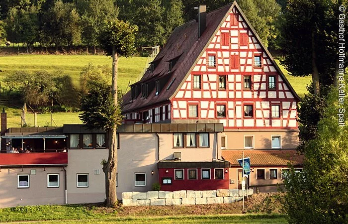 Ein großes vierstöckiges Fachwerkhaus mit roten Balken und weißer Fassade mit in einer grünen Landschaft. Links daneben eine große Steintereasse mit Sonnenschirmen.