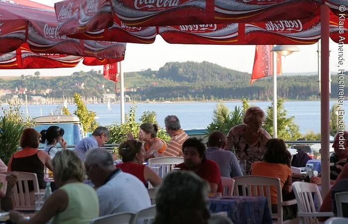 Gäste sitzen in einem Biergarten mit weißen Stühlen und roten Coca-Cola-Schirmen. Im Hintergund wird der Brombachsee von der Sonne beleuchtet.