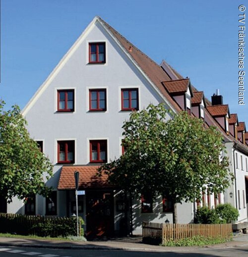 Die seitliche Ansicht eines großen vierstöckigen Hauses mit blauer Fassade und roten Fenstern. Direkt vor dem Gasthof stehen links und rechts vom Eingang Bäume.