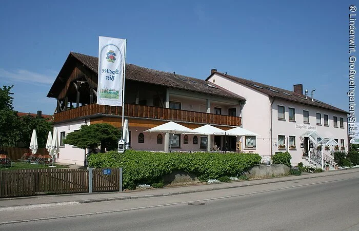 Ein großer Gebäudekomplex aus zwei großen langen Häuser mit jeweils zwei Stöcken. Zu sehen ist die Seitansicht entlang einer geteerten Straße. Die Fassade ist hellrosa. Das vordere Gebäude hat einen großen Holzbalkon rund um den 1. Stock.