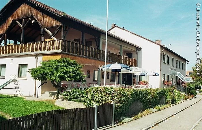 Ein Seitenansicht eines großen Gebäudekomplex aus zwei großen langen Häuser mit jeweils zwei Stöcken. Zu sehen ist die Seitansicht entlang einer geteerten Straße. Die Fassade ist weiß. Das vordere Gebäude hat einen großen Holzbalkon rund um den 1. Stock.