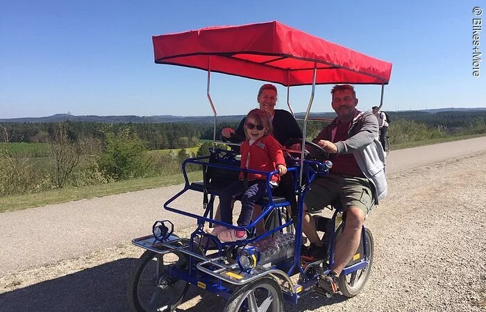 Eine Ehepaar mit Kind fährt auf einer Rikscha auf einem Schotterweg durch eine grüne Landschaft. Die Familie lächelt in die Kamera.