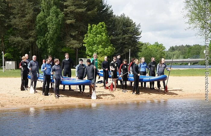 Zwei große Gruppen an Sportlern halten gemeinschaftlich jeweils ein großes Surfbrett. Die Bretter sind in etwa 2 Meter lang. Die Sportler tragen Neoprenanzüge und stehen an einem Strand mit Paddeln in der Hand.