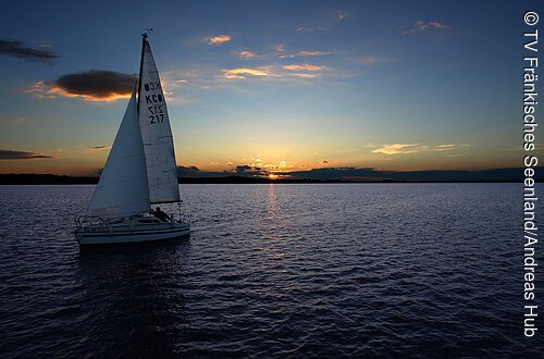Ein Segelschiff fährt Richtung Sonnenuntergang. Der Horizont ist orange und blau. Das Wasser des Sees dunkelblau bis schwarz.