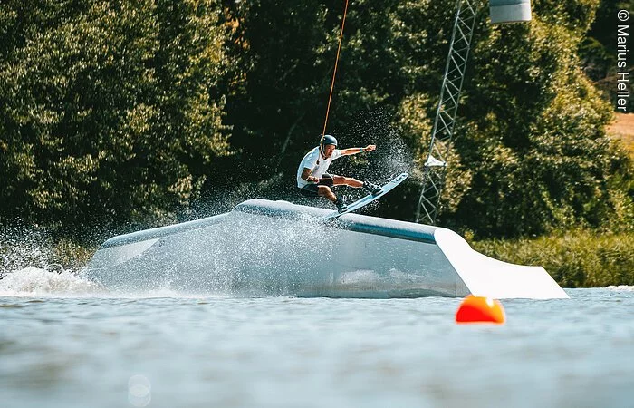 Ein Wakeboarder fährt über ein Hindernis im Wasser. Dahinter am Ufer grüne Bäume und Sträucher.