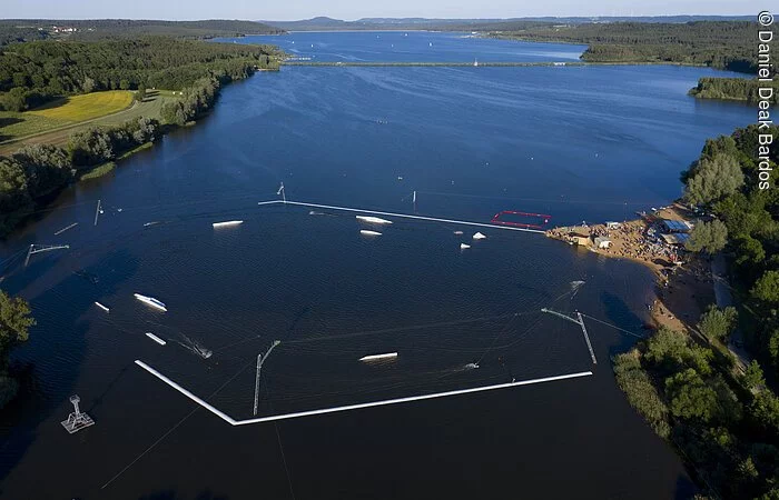 Luftbild von der Wakeboardanlage. Im See Hindernisse für die Wakeboardfahrer.  Am See ein Strand mit vielen Besuchern. Der See ist umgeben von grünen Bäumen und Sträuchern.