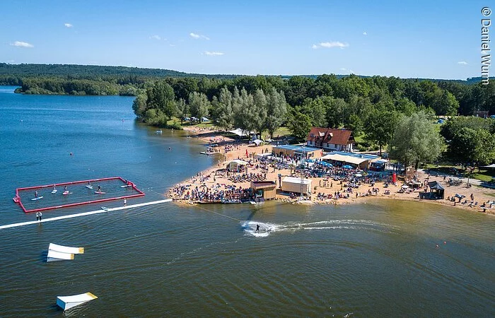 Luftbild von der Wakeboardanlage. Im See Hindernisse für die Wakeboardfahrer.  Am See ein Strand mit vielen Besuchern. Dahinter grüne Bäume und Sträucher.