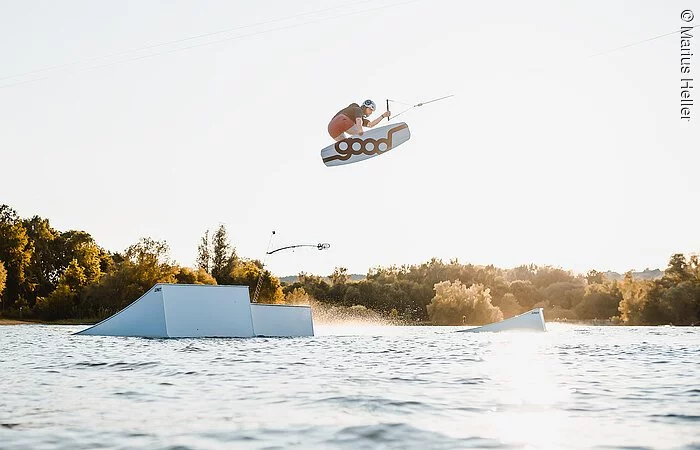 Ein Mann mit Wakeboard springt über ein Hinderniss im Wasser. Dahinter am Uder Bäume und Sträucher.