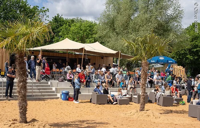 Eine Tribüne aus Stein dirket an einem Sandstrand. Darauf sitzen viele Menschen. Dahinter ein Sonnensegel und grüne Bäume.