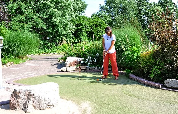 Ein Frau mit Sommerbekleidung steht auf einer grünen Minigolfbahn.  In den Händen hält sie einen Schläger und setzt gerade zum Schlag an. Im Hintergund der Minigolfbahn wachsen grüne Büsche und Sträucher.