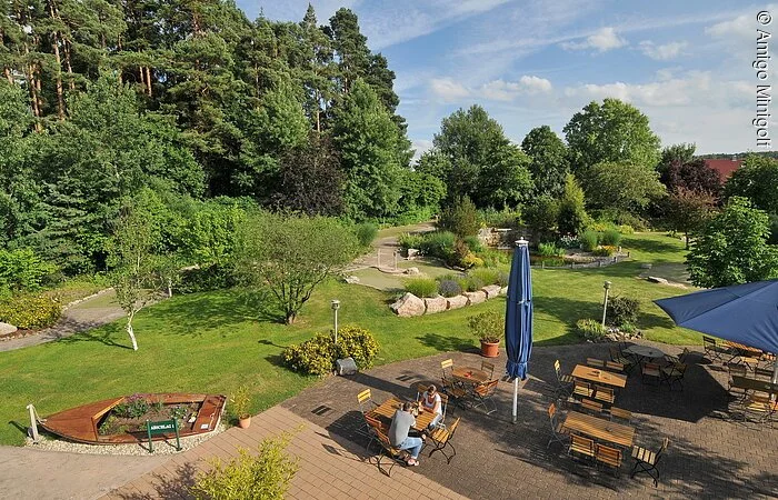 Kleiner Biergarten auf einem gepflasterterten Hof. Zwei Gäste sitzen an einem Tisch. HInter dem Biergarten eine grüne Wiese. Dahinter Bäume.