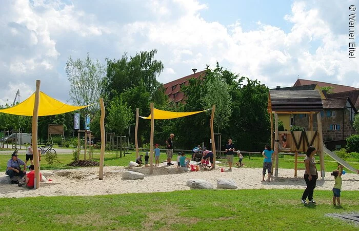 Ein Spielplatz auf Schotterfläche umgeben mit grüner Wiese. Zwei kleine gelbe Sonnensegel. Rechts daneben eine Rutsche. Kinder und Eltern spielen auf dem Spielplatz. Im Hintergund Büsche und Bäume.