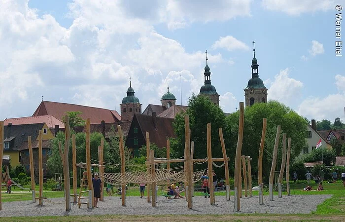 Im Vordergrund ein Spielplatz auf einer Schotterfläche umgeben von grüner Wiese. Auf dem Spielplatz ein Klettergerüst aus Holzpfählen, Plattformen und Netzen. Im Hintergund große Büsche und Bäume, dahinter Häuserdächer und drei Türme einer Kirche.
