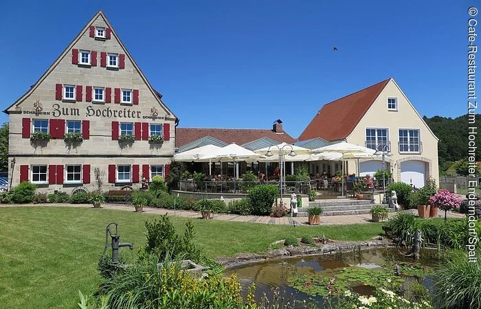 Ein großes fünfstöckiges Haus mit fränkischem Baustil mit roten Fensterläden. Rechts daneben eine große Gartenterasse mit weißen Sonnenschirmen. Recht daneben eine Garage mit zwei Toren. Vor dem Gebäudekomplex ein kleinerGartenteich auf einer großen grünen Wiese.