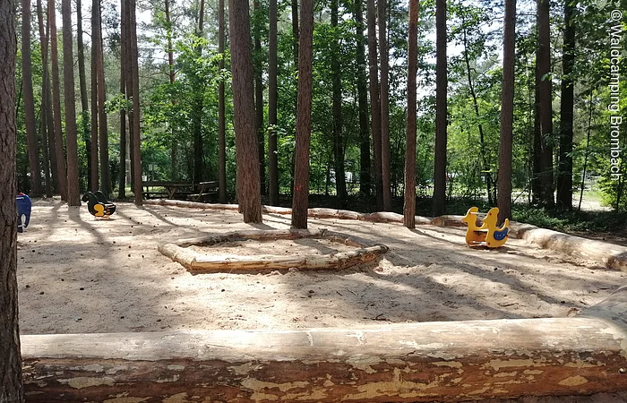 Spielplatz im Wald mit Sandboden, umgeben von Bäumen. Zwei Wipptiere sind im Sand zu sehen.