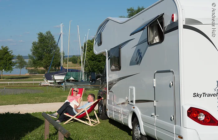 Eine Frau und ein Mann sitzen in zwei Liegestützen link neben einem Wohnmobil. Im Hintergund lagern Boote auf Anhängern. Dahinter beginnt der See.